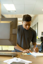 African-American student in a library, taking notes on paper by a computer monitor.