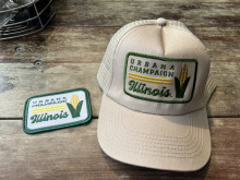 An iron-on patch and a tan baseball hat on a wood picnic table surface, both with an embroidered design featuring a large ear of corn and the words Urbana Champaign Illinois