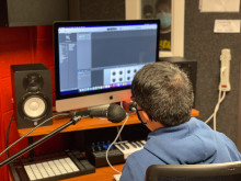 Shapiro Design Lab Winberg Media Production Room. There is a soundbooth in the background with a student inside speaking into a microphone and in the foreground another student sitting at a computer monitoring their recording. 