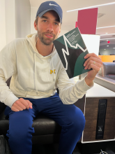 A Caucasian man wearing a blue cap with a white sweater holding a book.