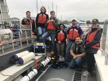 Seven scientist wearing orange life vest posing on the deck of a research ship with their sonar instrument. 