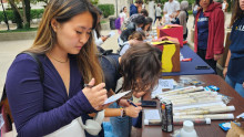 Students writing on postcards at a table outside (with trees in the background).