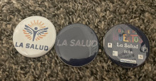 Picture showing 3 different pins, one in white that says la salud, one in black that says la salud, and one in black that says dia de los muertos la salud