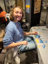 Student smiling at the camera with painted artwork on the table in front of her.