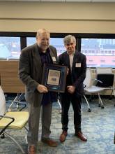 Ton Broos holding a framed copy of a Dutch pamphlet standing next to curator, Pablo Alvarez