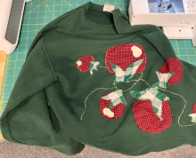 Image showing a dark green sweater on a green cutting mat. The sweater has tomatoes on a vine sewn onto it. The tomatoes are made out of 2 different plaid fabrics one red and one green.