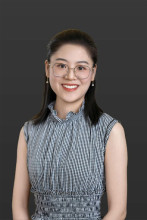 An Asian woman in a checkered dress and wire-rimmed glasses smiles in her headshot