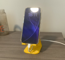 A charging Apple Iphone sitting on a yellow 3D printed phone stand. The stand is triangular; the back side has a hole for the charger, and the front side has two hooks to hold the phone while not blocking the port.