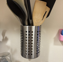 A metallic silver IKEA utensil holder hanging on a white wall. The holder is a tall cup with small holes perforating the sides, and it is holding an assortment of kitchen spatulas and utensils.