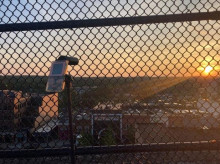 A Gameboy console, with an attached camera, standing on a tripod taking a picture of a sunset and city-scape from behind a chain-link fence.