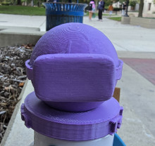 A blue 3D printed model of a sphere "wearing" a set of VR glasses across its middle. The sphere is on a blue 3D printed stand. The photo of the model was taken outside on a concrete ledge, so in the background can be seen a grass park, a blue trash can, pavement, and students talking.