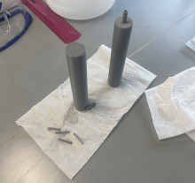 A set of gray 3D printed parts on a white napkin standing on a gray table. On the napkin are two tall cylinders, and one has a small piping end on the top. Additionally, there are four small tubes laying sideways on the end of the napkin.