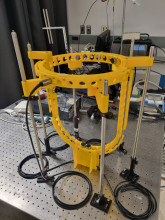 A yellow 3d printed prototype of a transducer unit holder supported by silver metal rods. The prototype has a square stand, which connects to a tall U shape, with a horizontal circle on top with 6 sleeves for the metal rods. Throughout the yellow plastic is one row of small holes to place transducer units.