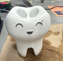 A white 3d printed toothbrush holder shaped like a tooth (molar). The holder has a smiling face in black outline on the front,and has 2 holes in the top to place toothbrushes.