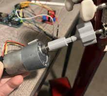 Gray 3d printed cylinder and gear attached to a guitar peg on the right side and a motor on the left side. The table behind the guitar is a wooden table with a lot of wires and circuit boards. 