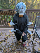 A child kneeling on a leaf covered patio. They are wearing black pants, a black hoodie, and red shoes. Along their arms are 4 gray hands with gold wrists, and their face is covered by one big hand.