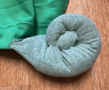 A roll of green fabric shaped to look like a chameleon tail. It is sewed onto the hem of a bright green shirt.