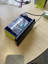 A blue 3d printed rectangular case with an open top - the case has sensors inside, which are connected by a wire to a battery pack strapped to the outside of the case. The (open) top of the case is taped over with scotch tape. 