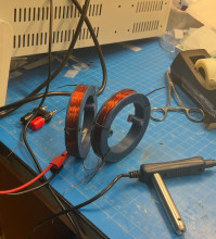 Two 3D printed gray bobbins, circular, on a workbench with copper wire coiled around them.