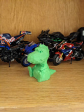A neon green, 3D printed toy dinosaur. The toy dino resembles a T-Rex, with a large snout and short arms, and it is sitting down with its legs in front and its tail behind it. The toy is sitting on a wooden shelf, with toy motorcycles behind it. 
