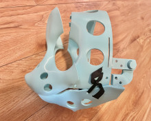 A light gray skeleton for a mask, laying sideways on a wooden table. there are holes for the eyes and mouth, and other smaller holes to attach the rest of the mask through.