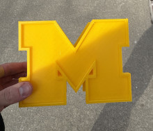 A yellow 3D printed block M pencil holder being held in front of a gray sidewalk. The block M used is the University of Michigan "M".