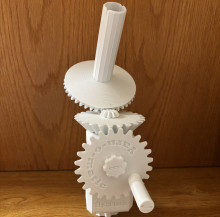 A white 3d printed mechanical yarn winder. There are three main gears on a rectangular base - the lowest gear has a handle for the user to spin up and down, which rotates the middle gear which connects to the gear on top of the mechanism, which has a tall rod sticking up from the middle to collect wound yarn.