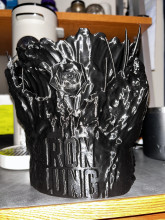 A black, tall 3D printed bucket placed on a wooden table. At the base of the bucket is the words "Iron Lung", and the top of the bucket is made to look like a fish's mouth with sharp, needle-like teeth.