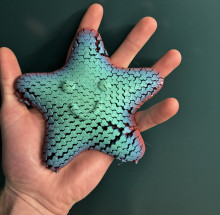 A multicolored 3D printed flexible starfish toy, held by an adult (white) hand in front of a dark green background. The toy is sea green in the middle, where it has a smiley face, spreading out towards the arms/edges it turns a light blue, and the edges of the arms are a muted, metallic red.