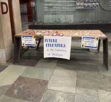 A photo of a table next to a column on a gray and green tiled floor. The table has a variety of candy and stickers on it, and hanging off the front of the table are three signs. There are two identical small signs on either end of the table: "Sticker and Candy sale! / Stickers: $1 / Candy: 2 for $1". The large sign in the middle is blue vinyl cut lettering on posterboard: " Future Educators at the University of Michigan".