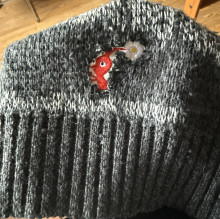 The hem of a sweater being held up to the camera. The sweater is dark gray with a light gray stripe on top of the dark grey ribbed knit. Above the light gray stripe is an embroidered cutout of a red pikmin (a small bipedal video game character with a white flower on its head)