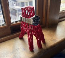 A 3d printed webbed red hand used as a stand to hold a silver watch. The hand is stood fingers down, wrist up, on a wooden windowsill.