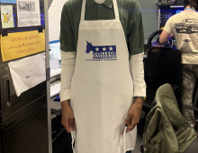 A photo of a dark skinned figure standing in front of a computer desk and chairs. The figure is wearing a white long sleeve shirt under a dark green short sleeved collared shirt. On top of that they are wearing a white apron that goes down to their knees, with a blue vinyl-cut logo of the University of Michigan College Democrats group. 