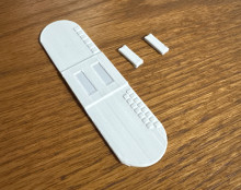 A 3d printed white rounded rectanglular strip. Along the right side is a strip of small squares, and in the middle of the rectangle is two small parallel rectangular cut-outs.