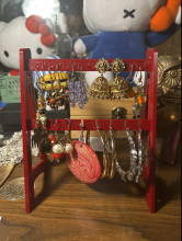 A red 3D printed earring stand on a wooden table with Hello Kitty dolls in the background. The stand is tall and uses A-stand legs to stay upright. There are two rungs to hold earrings on, and each rung holds about five pairs of hanging earrings or hoops