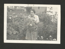 A light-skinned Black woman in glasses, wearing a dress, cardigan, and glasses, stands in a garden behind a rosebush