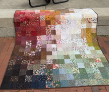A large patchwork quilt draped on brick pavement - the top of the quilt is placed on a taller concrete level. Each corner of the quilt is a different color: dark blues in the bottom left, grayish-greens in the bottom right, dark reds in the top left, and white/yellow in the top right. each section moves towards the middle and disperses in the center.