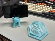 Two light blue 3D printed models on a gray desk in front of a computer keyboard. The model on the left is a phone holder shaped like a cat bending down or stretching. On the cat's back a phone is balanced sideways, where you can see the black screen. The model on the right is a hexagonal model of a mountain slope with lines detailing snowboard trails. There is writing on the base, but it is not legible in this photo.