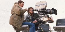 two men positioned around a film camera, one with eye to the lens