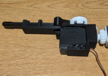 A black 3D printed holder for a servomotor. The holder has a squarish base to the right of the figure, with holes on the sides for the servomotor wires and white circular connectors and gears to go through. The holder also has a long finger-like piece extending out to the left.