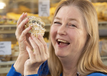 Head and shoulder photo of a blonde woman smiling and holding some kind of small baked good (maybe cake or cornbread?)