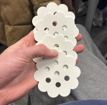 An image of a white hand holding a collection of four flat, white 3D printed flowers. Each flower has seven holes in the middle, shaped in a hexagonal pattern with one in the center. The flowers overlap each other, fanning down the hand (inward towards, towards the photographers body)