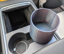 The center console of a car with a 3D printed cup holder in the right cup holder. The interior of the car is gray, and the image shows the two cup holders, and behind them (towards the front of the car) is a closeable hole. The 3D printed cup holder is tall, and wider than the original cup holder to be able to hold big bottles. It is printed in a shimmery blue-purple color.