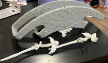 A pair of 3D printed white hairpins with a gray 3D printed traditional crown. Each hairpin is a long stick, meant to look like a stem, with a flower and buds at the end, and they are laid down across the image next to each other, with the left flower sitting on the end of the right stem. The crown is a large rectangular shape, with the edges of the rectangle curving down to look semicircular from the front; the top of the crown is much longer than the bottom. All the 3d prints are laying on a black table. 