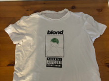 White t-shirt on a wooden surface, displaying a graphic with the word "blond" above a drawing of a person with green hair covering their face. The design includes a "Parental Advisory Explicit Content" warning beneath the illustration.