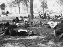 Black and white photograph of people lying on the grass among trees. Stone buildings in background. 