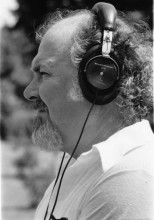 profile headshot of an older person with light skin, grey and white facial hair, and curly grey hair wearing headphones and a white collared shirt
