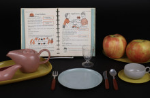 A complete setting of child sized dinnerware and flatware, complete with a small teapot and teacup is in the foreground, while in the background an opening of the Betty Crocker Cookbook for Boys and Girls is visible alongside two large apples. 