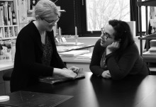 two women standing at a bench face each other in conversation as one works on a book