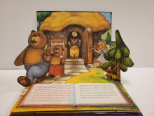 Pop-up page spread showing the three bears leaving on a walk, with a young blonde girl (Goldilocks) peeking out from behind a tree